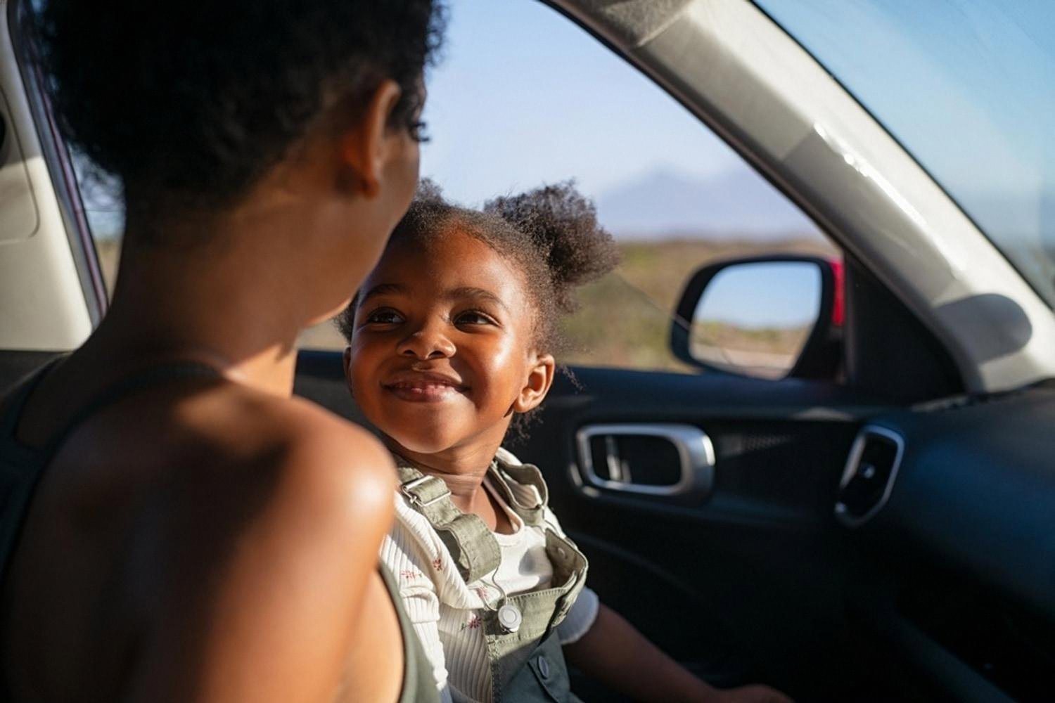 Mother and young daughter smiling at each other inside a car during a family trip abroad